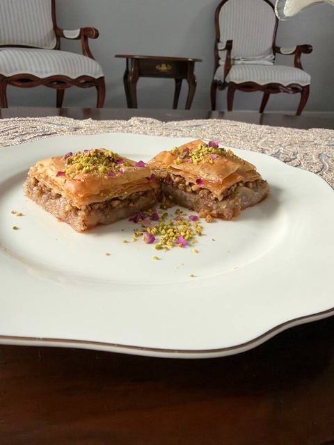 Two baklava squares topped with crushed pistachio on a white plate over a lace placemat